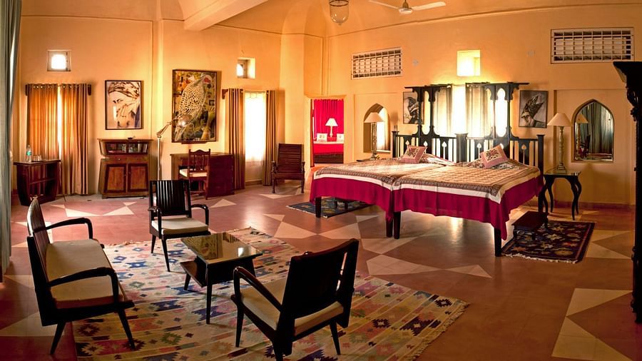 A large room is furnished with two beds covered with fabric, multiple seating chairs, tables, cabinets, and framed pictures on the walls inside Tijara Fort-Palace - 19th Century, Alwar.