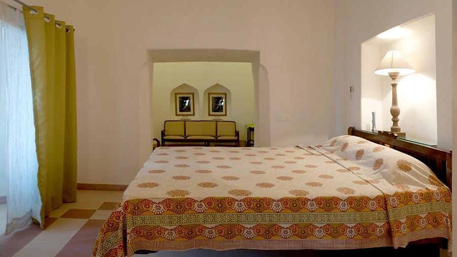 A bedroom features a bed with patterned bedding, a bedside table with a lamp, a window with curtains, and a view into an adjacent room with a sofa and framed pictures at Tijara Fort-Palace - 19th Century, Alwar.