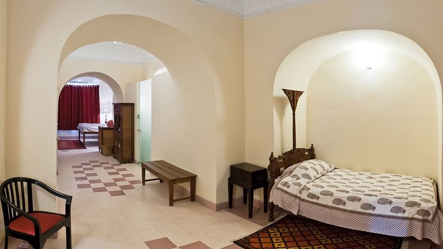 A bedroom with a bed, a floor lamp, a side table, a chair, and a bench is visible, with an arched doorway leading to another room with a bed and furniture at Tijara Fort-Palace - 19th Century, Alwar.