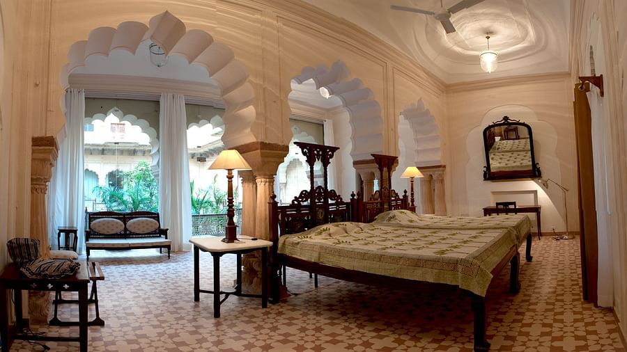 A bedroom features two beds with light coverings, bedside tables with lamps, arched doorways, and a seating area with a bench near a window at Tijara Fort-Palace - 19th Century, Alwar.