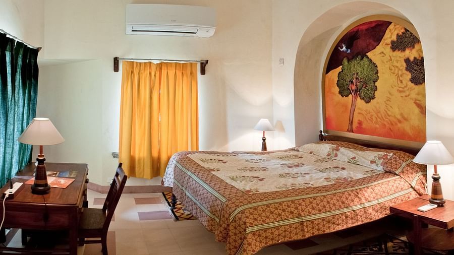 A bedroom features a bed with patterned bedding, two bedside tables with lamps, a writing desk with a chair, and two windows with curtains inside Tijara Fort-Palace - 19th Century, Alwar.