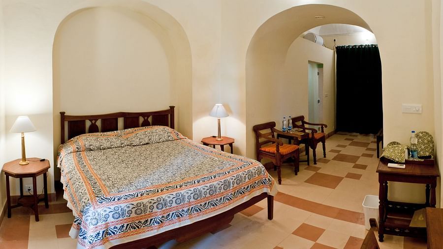 A bedroom contains a bed with patterned linen, two round side tables with lamps, and a view into an adjacent area with a table and chairs at Tijara Fort-Palace - 19th Century, Alwar.