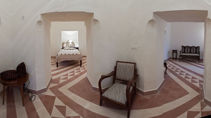 A room with tiled flooring has arched doorways leading to other areas, one with a bed and another with a bench, along with a chair and a small table in the foreground at Tijara Fort-Palace - 19th Century, Alwar.