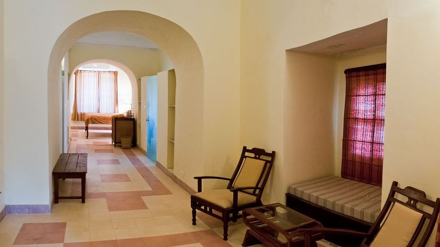 A hallway at Neemrana's Tijara Fort Palace features arched openings, tiled flooring, a bench, chairs, a coffee table, and a window seat with patterned cushions and a dark curtain.