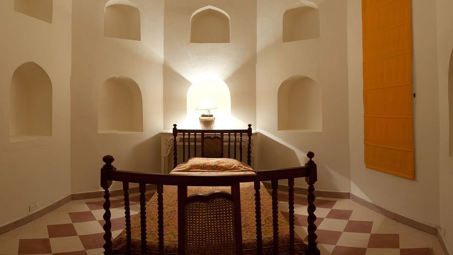 A bedroom contains a bed with bedding, a light source behind the headboard, and arched niches in the walls inside Tijara Fort-Palace - 19th Century, Alwar.