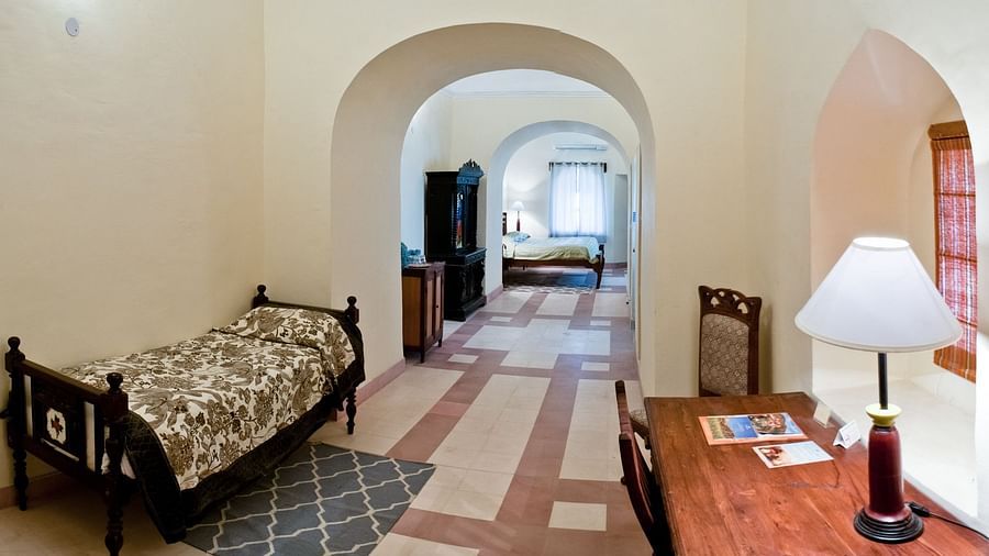 A suite at Neemrana's Tijara Fort Palace features a bedroom with a single bed and patterned linens, and a study area with a desk and chair, all connected by a tiled hallway with arched doorways and a wardrobe.