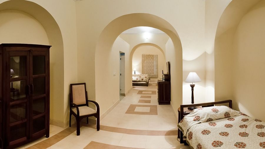 A hallway at Neemrana's Tijara Fort Palace features arched openings, tiled flooring, a wardrobe, a chair, and a glimpse into a guest room with a single bed and a floor lamp.