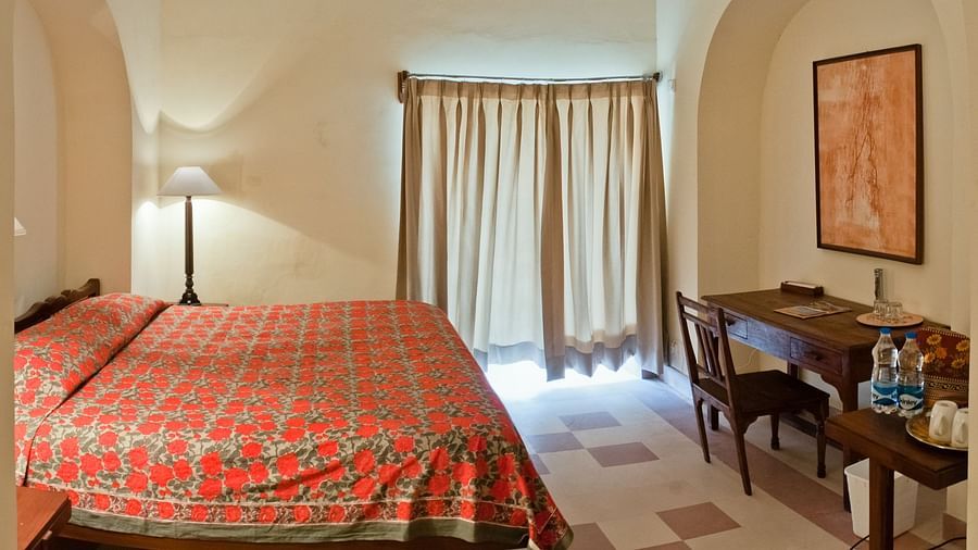 A guest room at Neemrana's Tijara Fort Palace features a bed with a red floral cover, a floor lamp, a window with beige curtains, a desk with chairs, framed artwork on the wall, and tiled flooring.