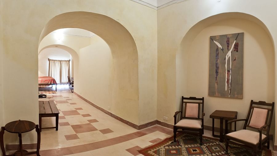 A hallway at Neemrana's Tijara Fort Palace features arched openings, tiled flooring, a bench, a small table, and a seating area with two chairs and a small table in an arched alcove with artwork and a glimpse into a guest room is visible.