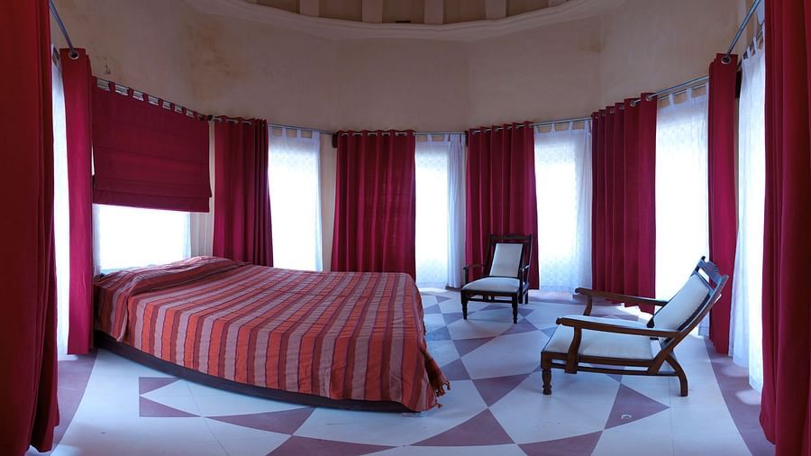 A round room includes a bed with striped bedding, multiple windows with curtains, and two chairs inside Tijara Fort-Palace - 19th Century, Alwar.
