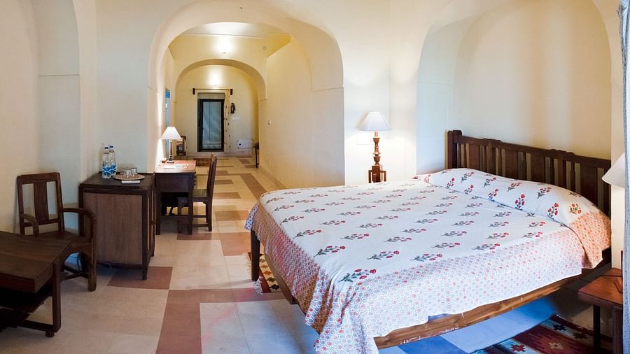 A suite at Tijara Fort-Palace - 19th Century, Alwar features a bedroom with a bed with white floral linens, a bedside table with a lamp, and an arched opening leading to a study area with a desk and chairs.
