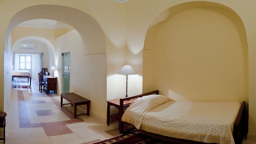 A guest room at Tijara Fort-Palace - 19th Century, Alwar features a single bed with white linens, a bedside table with a lamp, and an arched opening leading to a tiled hallway with a bench and a glimpse of a study area.