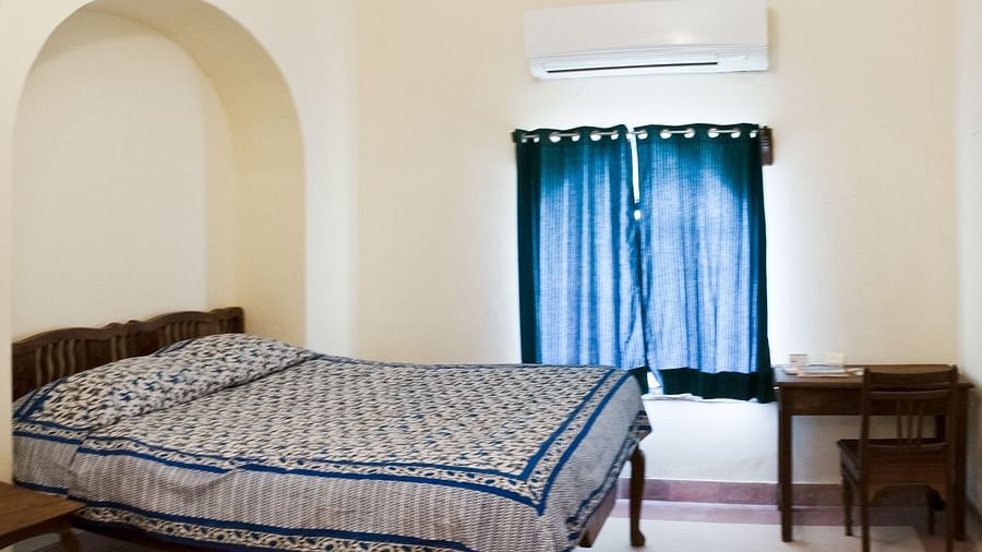A guest room at Tijara Fort-Palace - 19th Century, Alwar features a bed with blue and white patterned linens, a bedside table, a desk with a chair, a window with blue curtains, an arched niche in the wall, and air conditioning.