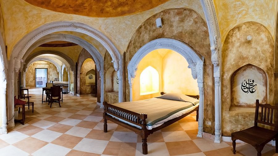 A unique guest room at Tijara Fort-Palace - 19th Century, Alwar features textured walls, arched doorways, a bed with a simple cover, a chair, and a window with an arched top.Tijara Fort-Palace - 19th Century_ Alwar Alwar Kotwara Mahal Facade_Tijara Fort Pa