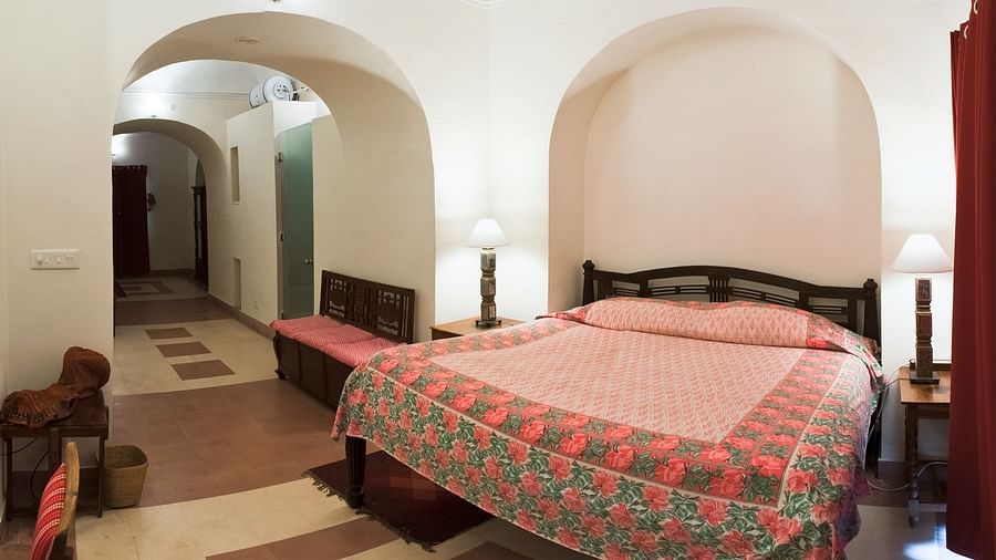 A guest room at Tijara Fort-Palace - 19th Century, Alwar features a bed with a pink and green floral cover, bedside tables with lamps, and an arched opening leading to a tiled hallway with a bench.