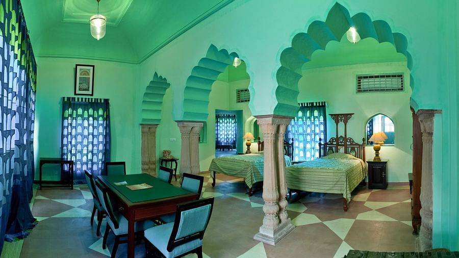 A room with multiple beds, bedside tables with lamps, a dining table with chairs, arched doorways, and windows with curtains is visible inside Tijara Fort-Palace - 19th Century, Alwar.