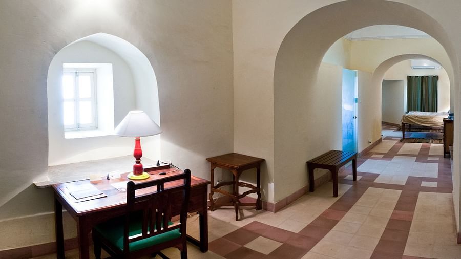 A study area at Tijara Fort-Palace - 19th Century, Alwar features a desk with a lamp and chair in an arched window alcove, a small table, and a view into a tiled hallway with a bench and a glimpse of a bedroom.