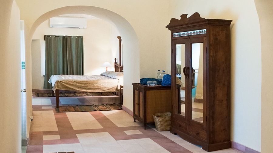 A common area at Neemrana's Tijara Fort Palace features arched doorways, tiled flooring, a mirrored wardrobe, a small cabinet, and a glimpse into a guest room with a bed and curtains.