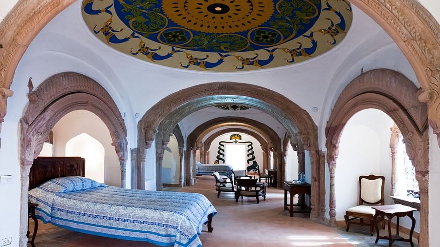 A suite at Tijara Fort-Palace - 19th Century, Alwar boasts a stunning painted ceiling, arched doorways, a bed with blue and white striped linens, a dining area with a table and chairs, and a seating area with a chair and small table.