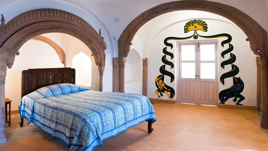 A unique guest room at Tijara Fort-Palace - 19th Century, Alwar features a bed with blue and white striped linens, arched doorways, and distinctive wall paintings in an arched alcove.