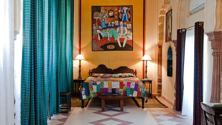 A bedroom features a bed with a patterned cover, two bedside tables with lamps, a painting on the wall, and windows with curtains inside Tijara Fort-Palace - 19th Century, Alwar.