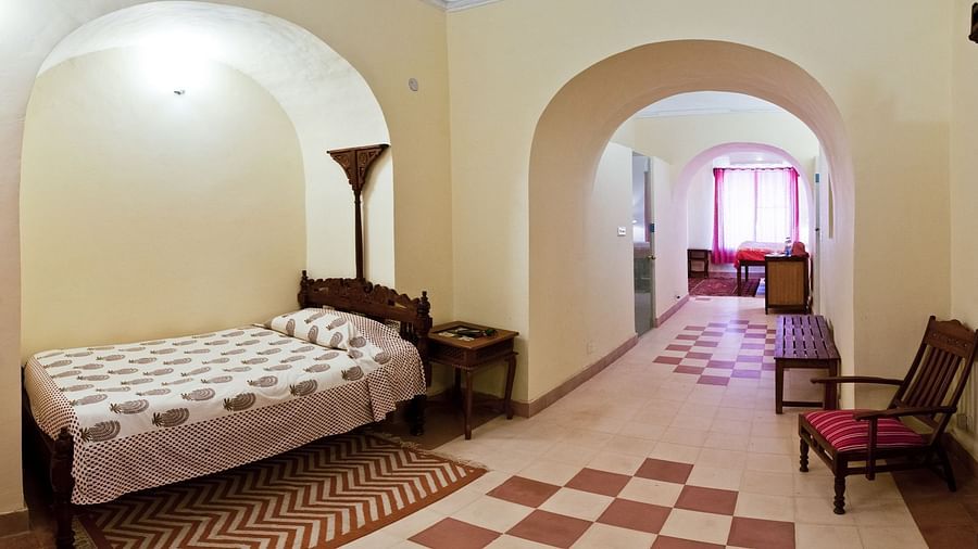A guest room at Neemrana's Tijara Fort Palace features a single bed with patterned linens, a bedside table with a lamp, a floor lamp, and an arched opening leading to a tiled hallway with a bench and chairs.