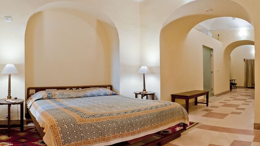 A guest room at Neemrana's Tijara Fort Palace features a bed with patterned linens, bedside tables with lamps, and an arched opening leading to a tiled hallway with a bench.