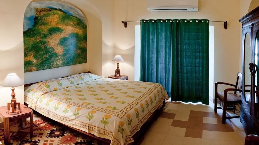 A bedroom features a bed with patterned bedding, two bedside tables with lamps, a window with curtains, an armchair, and a wardrobe inside Tijara Fort-Palace - 19th Century, Alwar.