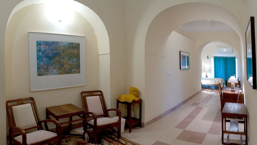 A hallway at Tijara Fort-Palace - 19th Century, Alwar features arched openings, tiled flooring, chairs, small tables, framed artwork on the walls, and a glimpse into a guest room with blue curtains.
