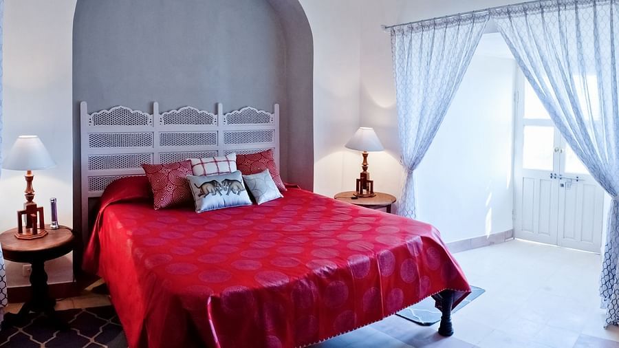 A guest room at Tijara Fort-Palace - 19th Century, Alwar features a bed with a vibrant red cover and decorative pillows, bedside tables with lamps, a white decorative headboard, an arched alcove, and a window with sheer white curtains.