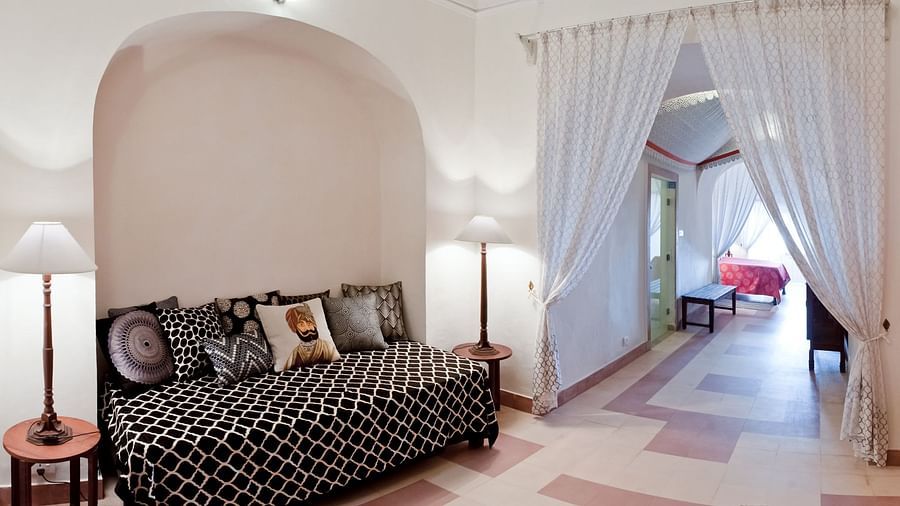A suite at Neemrana's Tijara Fort Palace features a seating area with a patterned daybed and decorative pillows, bedside tables with lamps, and an arched opening with sheer white curtains leading to another area.