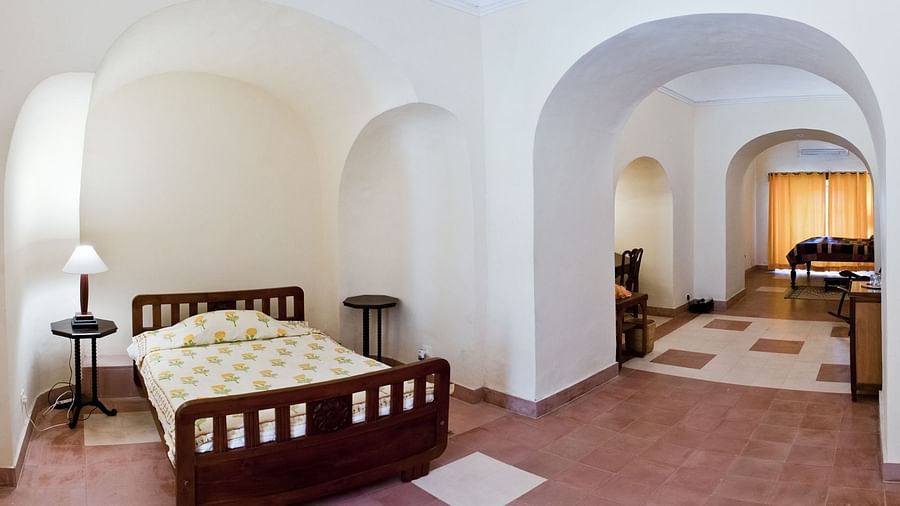A guest room at Tijara Fort-Palace - 19th Century, Alwar features a single bed with a yellow patterned cover, bedside tables with lamps, and an arched opening leading to a tiled hallway with a glimpse of another room.
