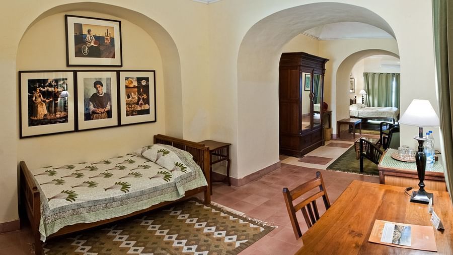A suite at Neemrana's Tijara Fort Palace features a bedroom with a single bed, framed artwork in an arched alcove, and a bedside table, as well as a study area with a desk, chair, and a glimpse of a dining space with a table and chairs.