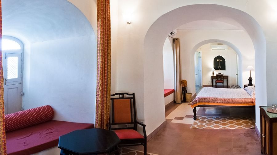 A suite at Neemrana's Tijara Fort Palace features arched doorways, a living area with a red daybed, a black table, and a chair, and a bedroom with a bed and patterned flooring.