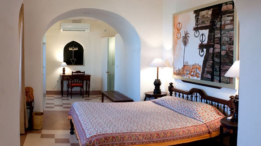 A suite at Tijara Fort-Palace - 19th Century, Alwar features a bedroom with a bed with patterned linens, bedside tables with lamps, artwork, and an arched opening leading to a study area with a desk and chair, and a bench.