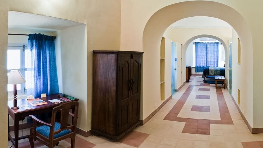 A hallway at Tijara Fort-Palace - 19th Century, Alwar features arched openings, tiled flooring, a wardrobe, and a glimpse into a study area with a desk and blue curtains, as well as a seating area with blue furniture.
