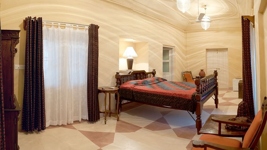 A bedroom is furnished with a bed covered in striped fabric, a bedside table, a window with curtains, and a chair near a wardrobe inside Tijara Fort-Palace - 19th Century, Alwar.