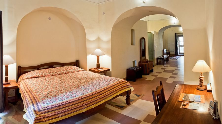 A guest room at Neemrana's Tijara Fort Palace features a bed with patterned linens, bedside tables with lamps, a desk with a lamp and chair, and an arched opening leading to a tiled hallway.