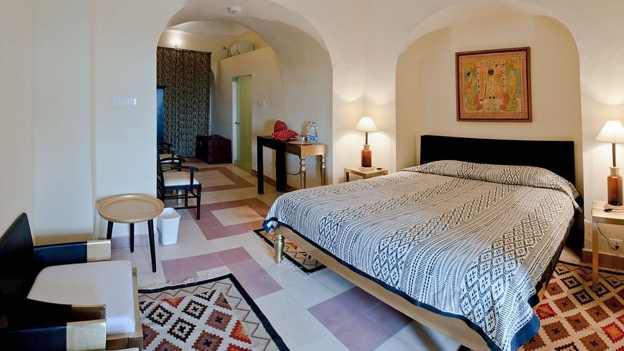 A suite at Tijara Fort-Palace - 19th Century, Alwar features a bedroom with a bed, bedside tables with lamps, artwork in an arched alcove, and a sitting area with chairs and a table, all with uniquely patterned flooring and arched doorways.
