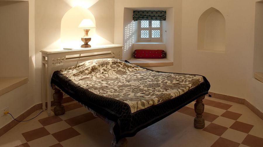 A bedroom features a bed with a patterned cover, a bedside table with a lamp, and a window with a curtain inside Tijara Fort-Palace - 19th Century, Alwar.