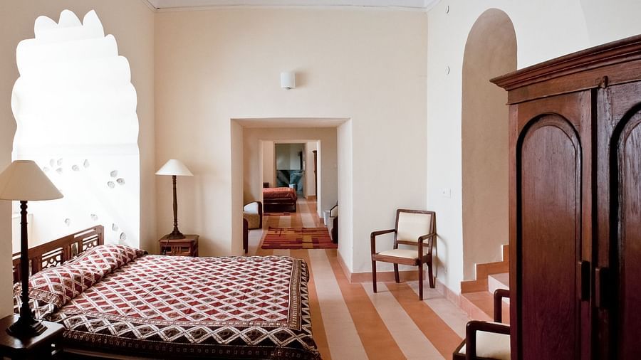 A guest room at Tijara Fort-Palace - 19th Century, Alwar features a bed with red and white patterned linens, bedside lamps in a decorative arched window alcove, chairs, a wardrobe, and a view into a hallway with tiled flooring.