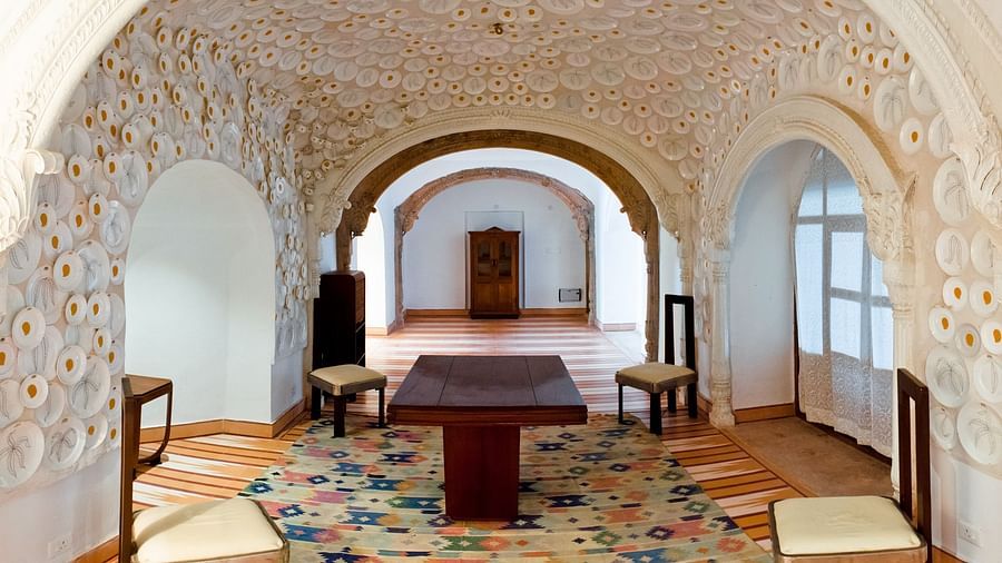 A beautifully decorated hallway at Tijara Fort-Palace - 19th Century, Alwar showcases intricate wall and ceiling carvings, arched openings, a dining table with chairs on a colourful rug, and a glimpse of a wardrobe in the distance.