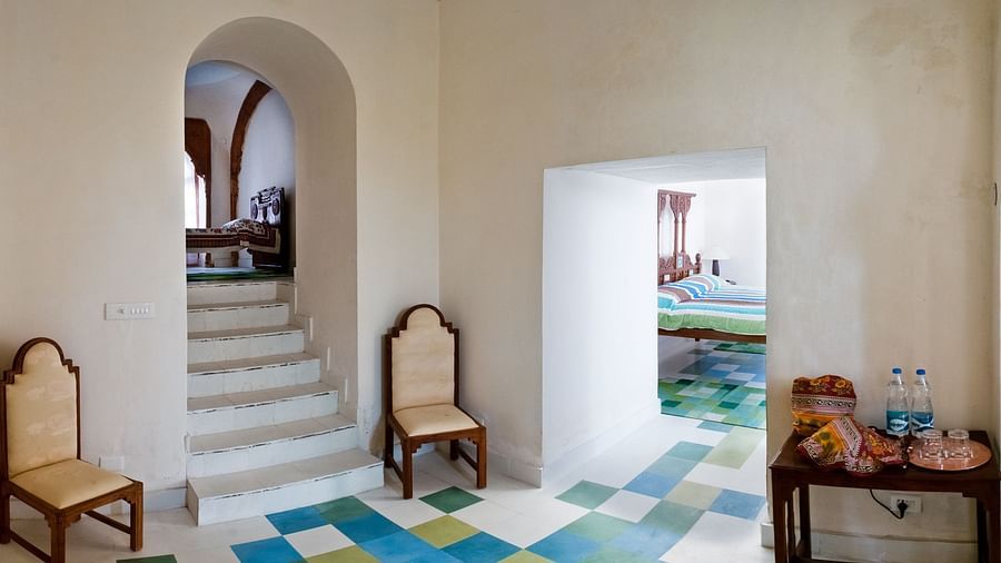 A common area at Tijara Fort-Palace - 19th Century, Alwar features arched doorways, tiled flooring with a blue and green pattern, chairs, a table with snacks and water bottles, and a glimpse of stairs and a bedroom.