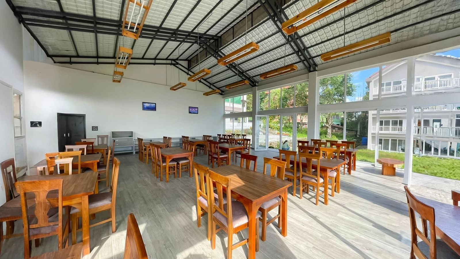 A spacious and bright dining area at Yashshree Resorts & Spa, Kalimpong, featuring numerous wooden tables and chairs, under a high ceiling with exposed beams.