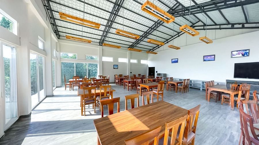A spacious and bright dining area at Yashshree Resorts & Spa, Kalimpong, featuring numerous wooden tables and chairs, under a high ceiling with exposed beams.