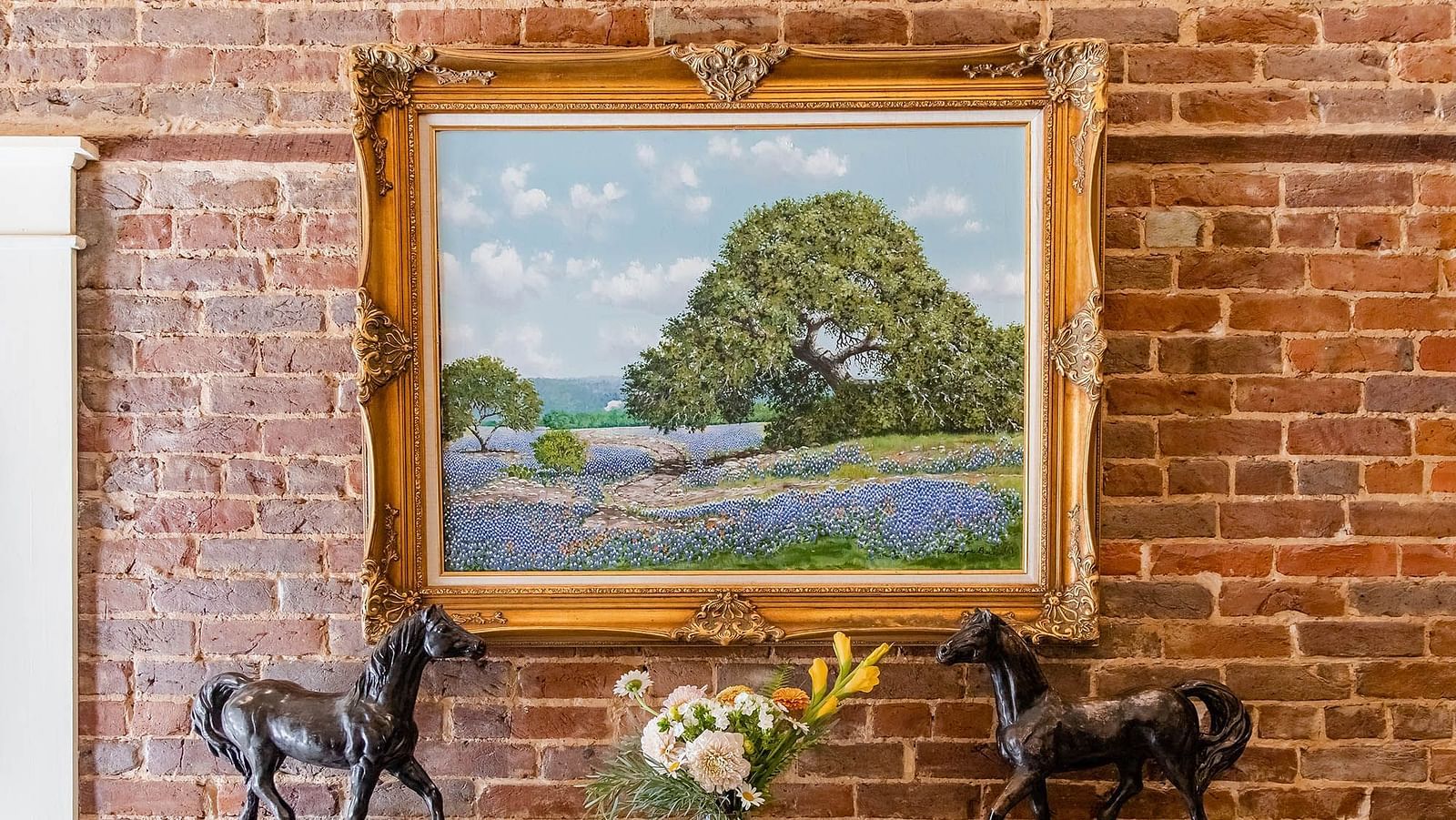 A framed landscape painting on a brick wall, with two horse statues on a wooden console table.- The Grand Hotel & Ballroom, McKinney