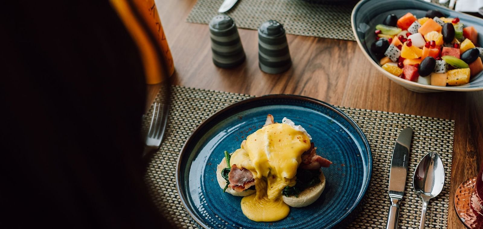 Close-up of Eggs Benedict with hollandaise sauce on a blue plate, accompanied by a bowl of mixed fruit salad.