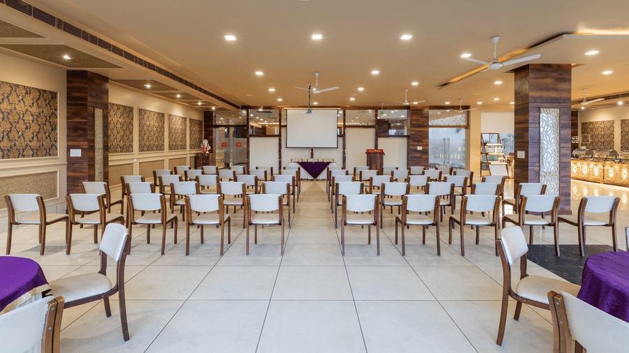 Hotel Highway Prince Paota banquet hall in theater-style setup with white chairs, purple accent tables, projector screens, and recessed lighting.