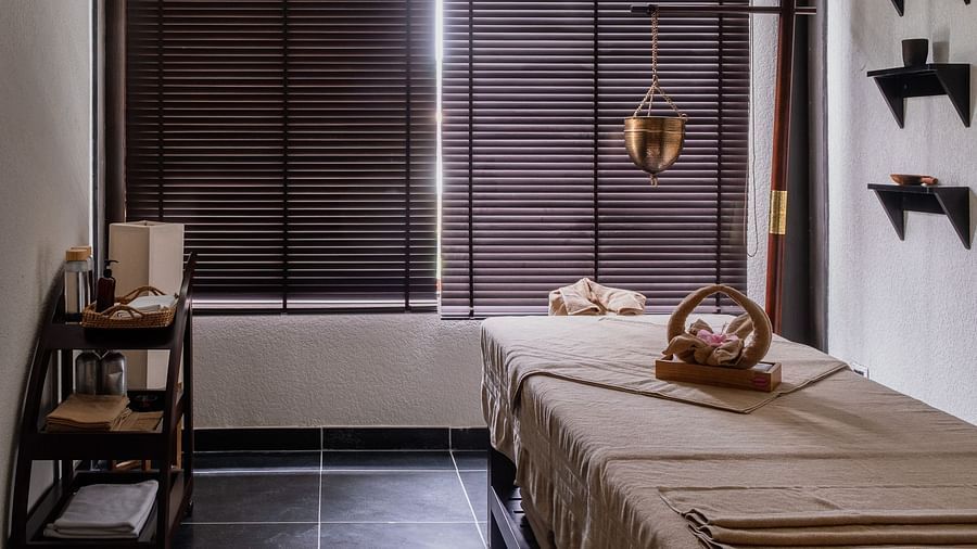A tranquil spa room with a massage table covered in a light-colored blanket, with a towel arranged as a swan on top. A hanging brass lamp, a wooden shelf with spa products, and a large window with wooden blinds create a serene atmosphere. This is an interi