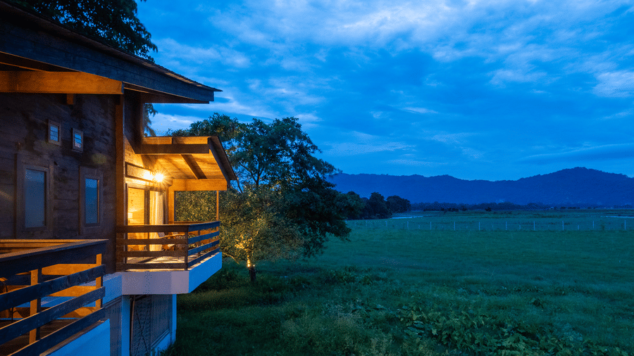 A view from a wooden balcony at Rhino & River Wildlife Retreat & Spa, Pobitora, looking out over a vast green field toward distant mountains under a deep blue twilight sky.